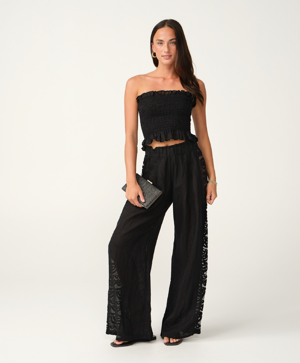 A woman modeling a black strapless cropped top and matching wide-leg pants with lace side panels, photographed against a clean off-white studio background.