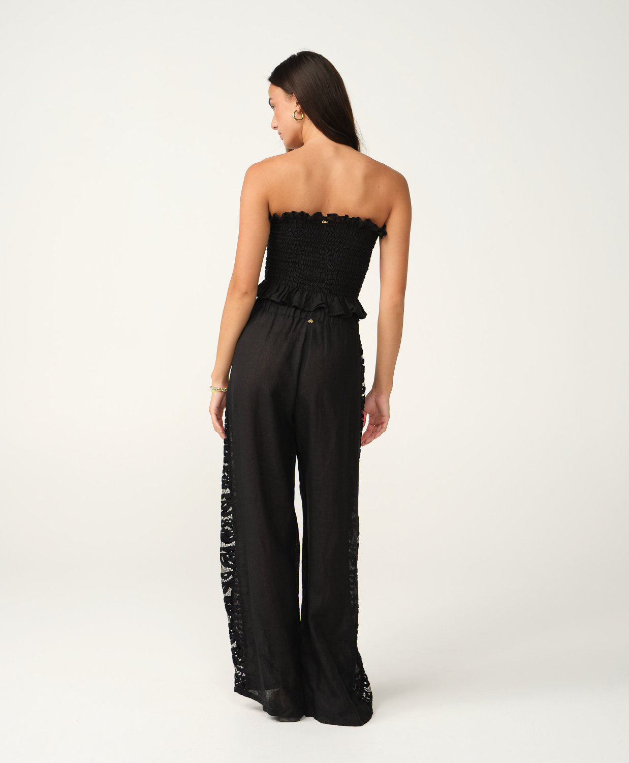 A woman modeling a black strapless cropped top and matching wide-leg pants with lace side panels, photographed against a clean off-white studio background.