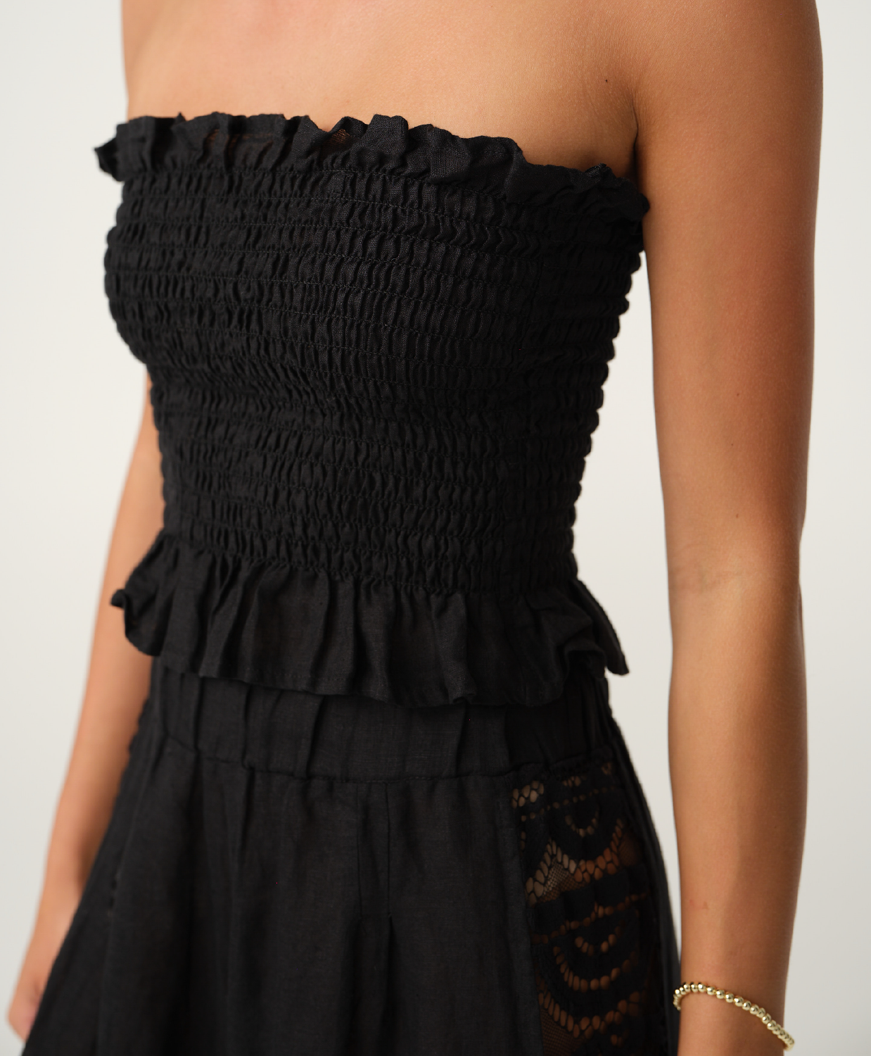 Close-up of a black strapless smocked crop top & pants with a ruffled waistband and side lace panel, worn by a model against a light neutral background.