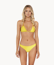 Blonde woman wearing a yellow ruched triangle top bikini standing in front of white wall.