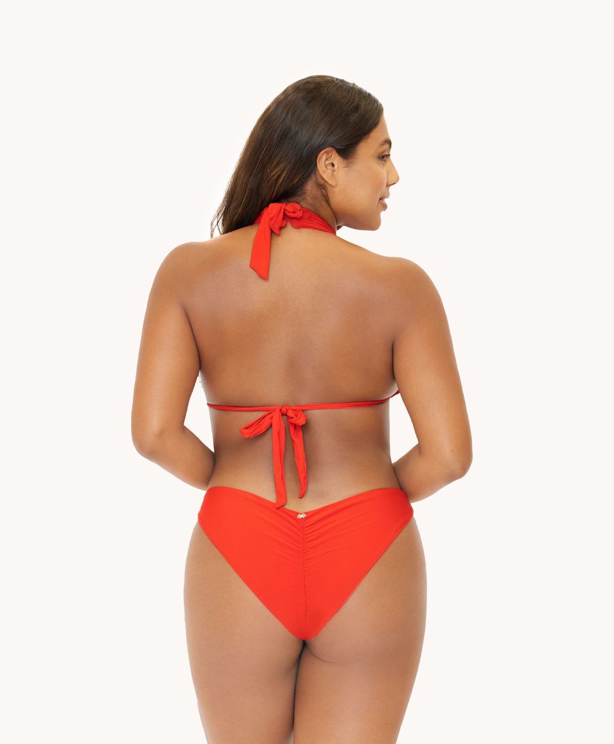 Back view of brunette woman wearing a neon orange bikini in front of a white background. 