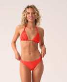 A woman wearing a red macrame triangle top bikini standing against a white wall.