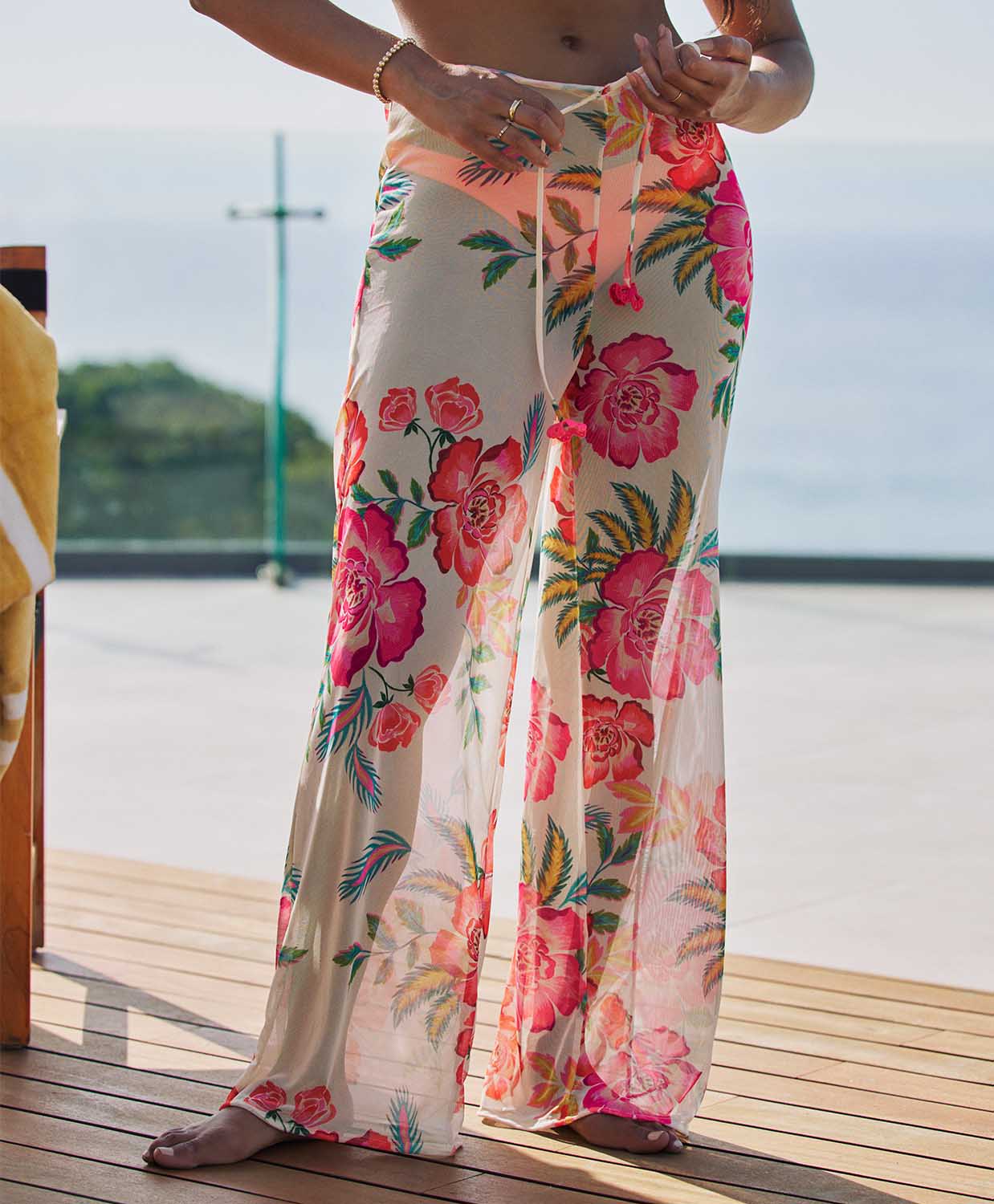 A woman wearing white pants with a floral print standing on a deck outside.