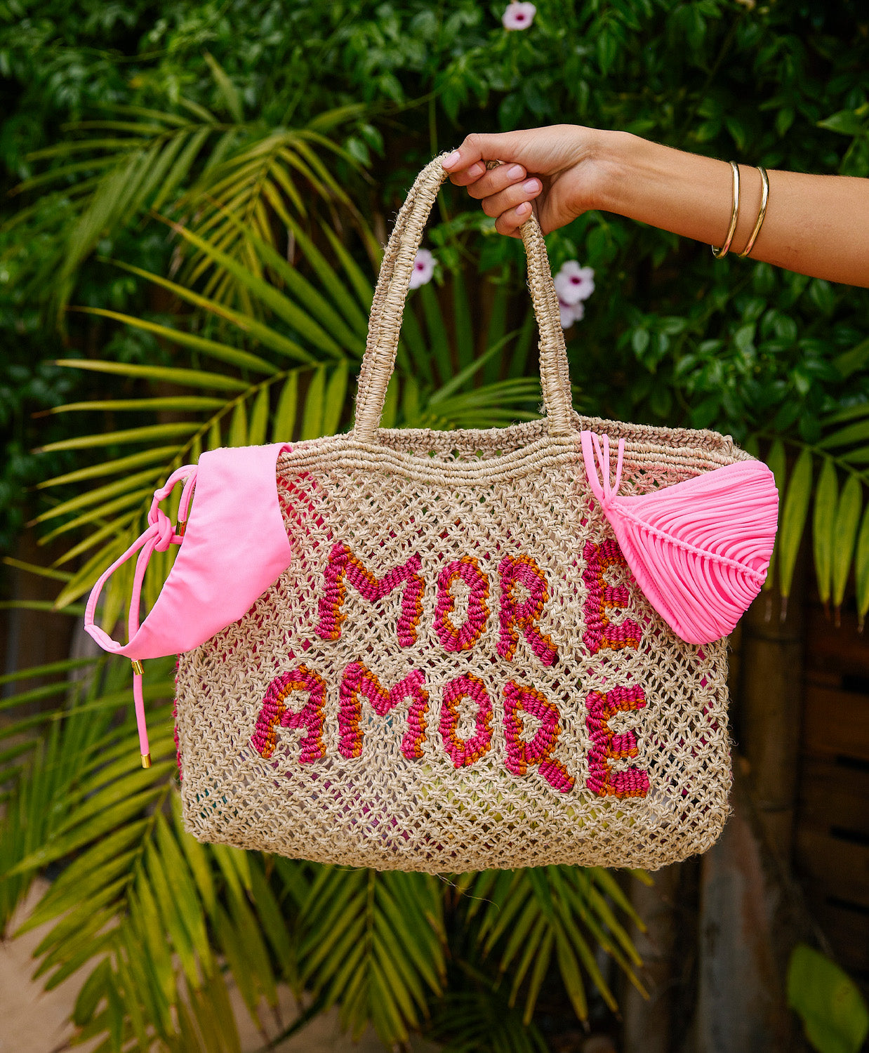 Woven bag with 'MORE AMORE' text and pink accents held by a person against a green leafy background