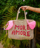 Woven bag with 'MORE AMORE' text and pink accents held by a person against a green leafy background