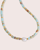 A multi colored necklace with a white pearl against a white wall.