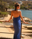 A woman stands on a balcony overlooking a beach and blue water, wearing a sleeveless blue maxi dress with a halter neck and an open back.