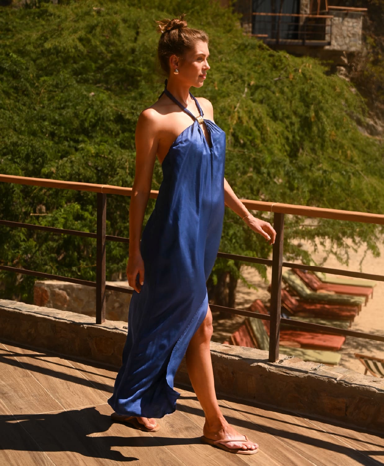 A woman wearing a blue halter-neck maxi dress walks along a wooden boardwalk outdoors with greenery and beach loungers in the background.