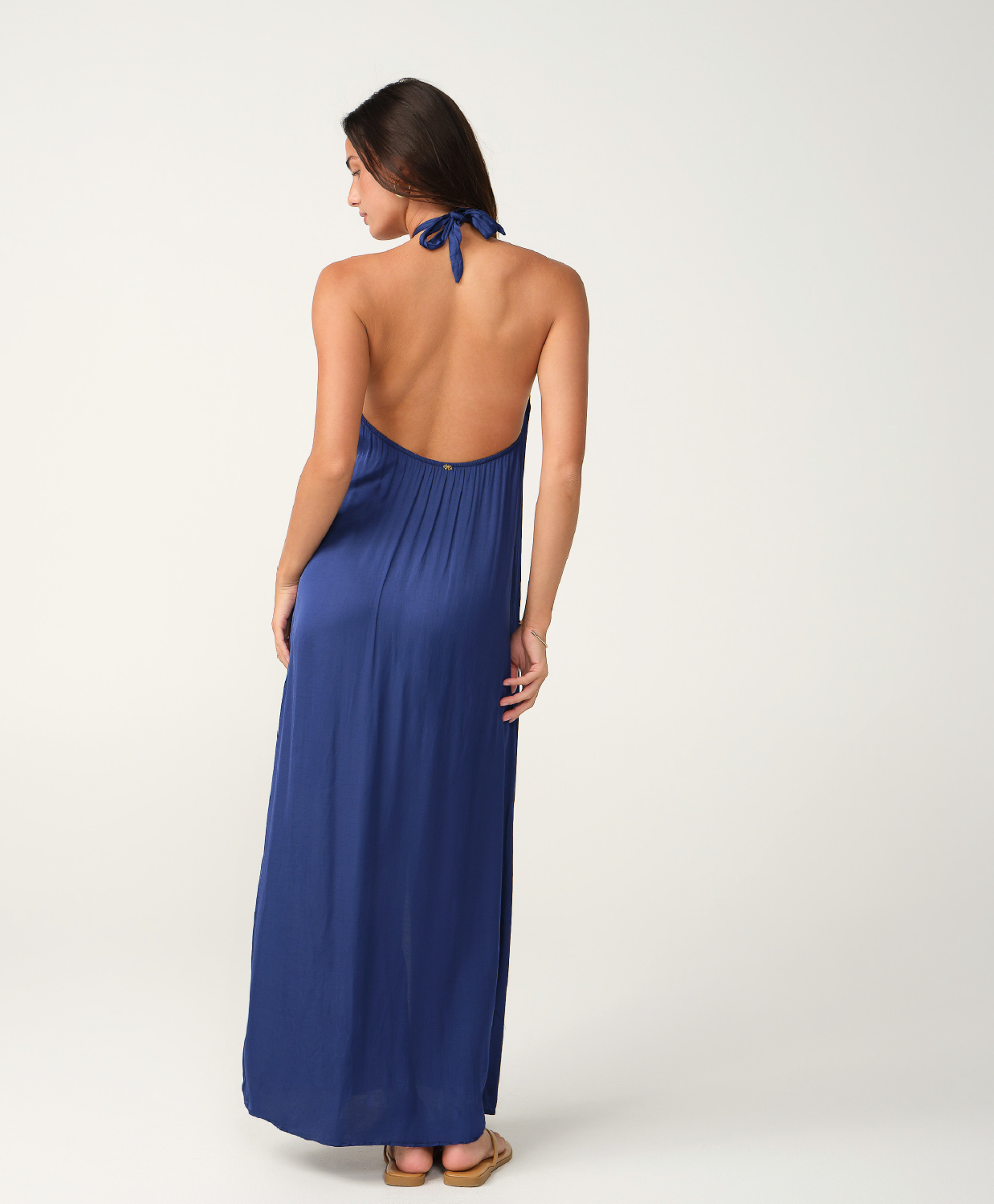 A woman modeling a blue halter-neck maxi dress from the back, featuring an open back and tie detail at the neck, set against a plain light gray studio background.