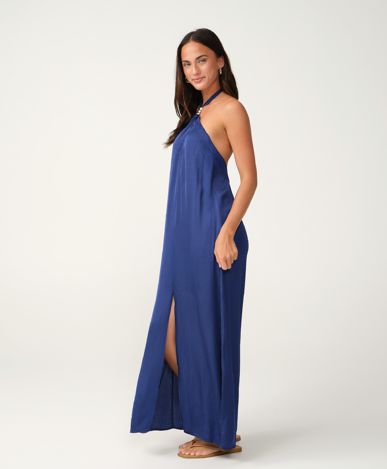A woman modeling a sleeveless, halter-neck maxi dress in a vibrant royal blue. The dress features a high thigh slit and is styled with tan flat sandals against a clean, light gray studio background.