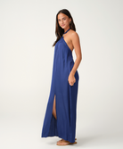 A woman modeling a sleeveless, halter-neck maxi dress in a vibrant royal blue. The dress features a high thigh slit and is styled with tan flat sandals against a clean, light gray studio background.