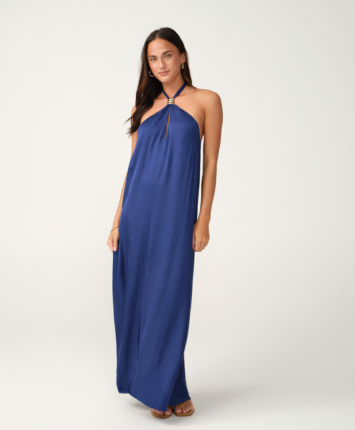 A woman modeling a royal blue halter-neck maxi dress with a decorative silver-toned embellishment at the neckline, standing against a plain light gray studio background.