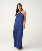 A woman modeling a royal blue halter-neck maxi dress with a decorative silver-toned embellishment at the neckline, standing against a plain light gray studio background.