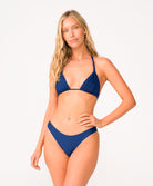 Blonde woman wearing a navy blue bikini in front of a white background. 