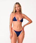 A woman wearing a navy triangle top bikini standing against a white wall.