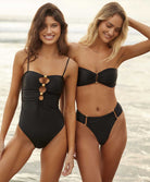 Two woman standing near the ocean. One woman wearing a black one piece swimsuit and the other is wearing a black bandeau bikini. 