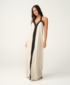 A woman modeling a long cream linen maxi dress with black trim and a black front placket, photographed against a seamless white studio background.