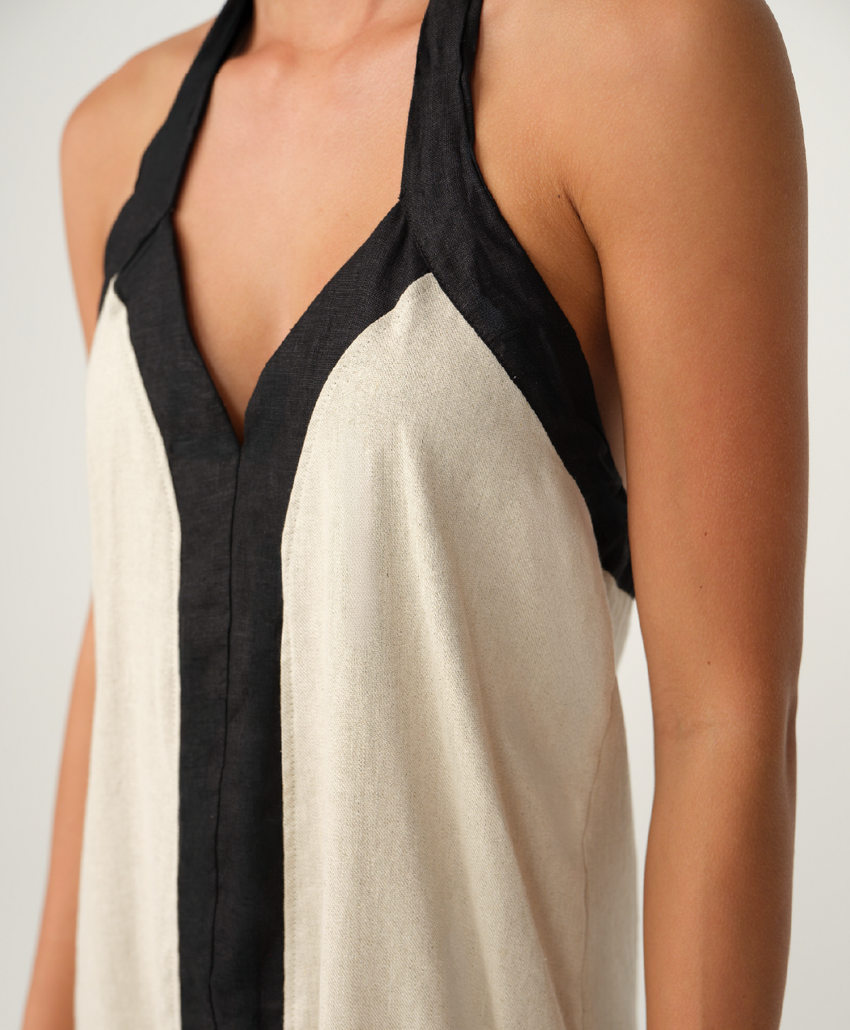 Close-up of a woman wearing a sleeveless halter-style top or dress in off-white with black trim and shoulder panels, against a plain light background.