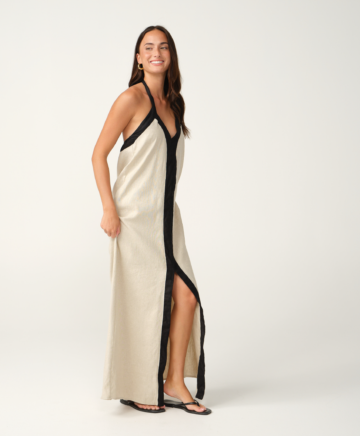 A woman modeling a long cream linen maxi dress with black trim and a black front placket, photographed against a seamless white studio background.