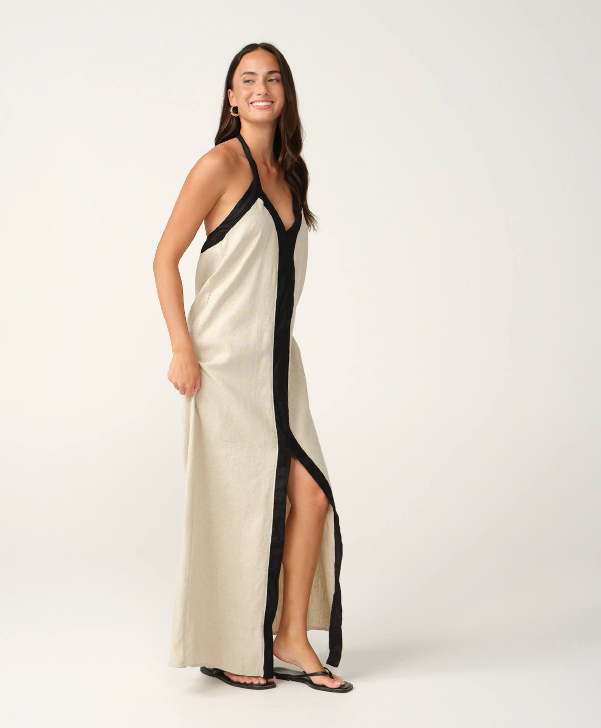 A woman modeling a long cream linen maxi dress with black trim and a black front placket, photographed against a seamless white studio background.