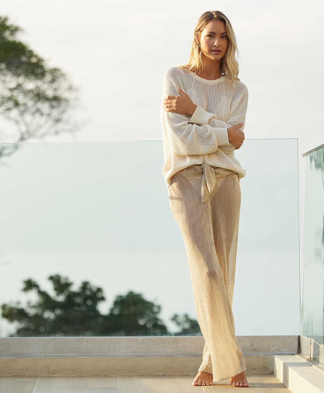 A blonde woman wearing an ivory knit sweater and long gold pants standing on a patio outside. 