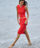 A woman wearing a matching red crochet top and shirt walking through water on a beach.