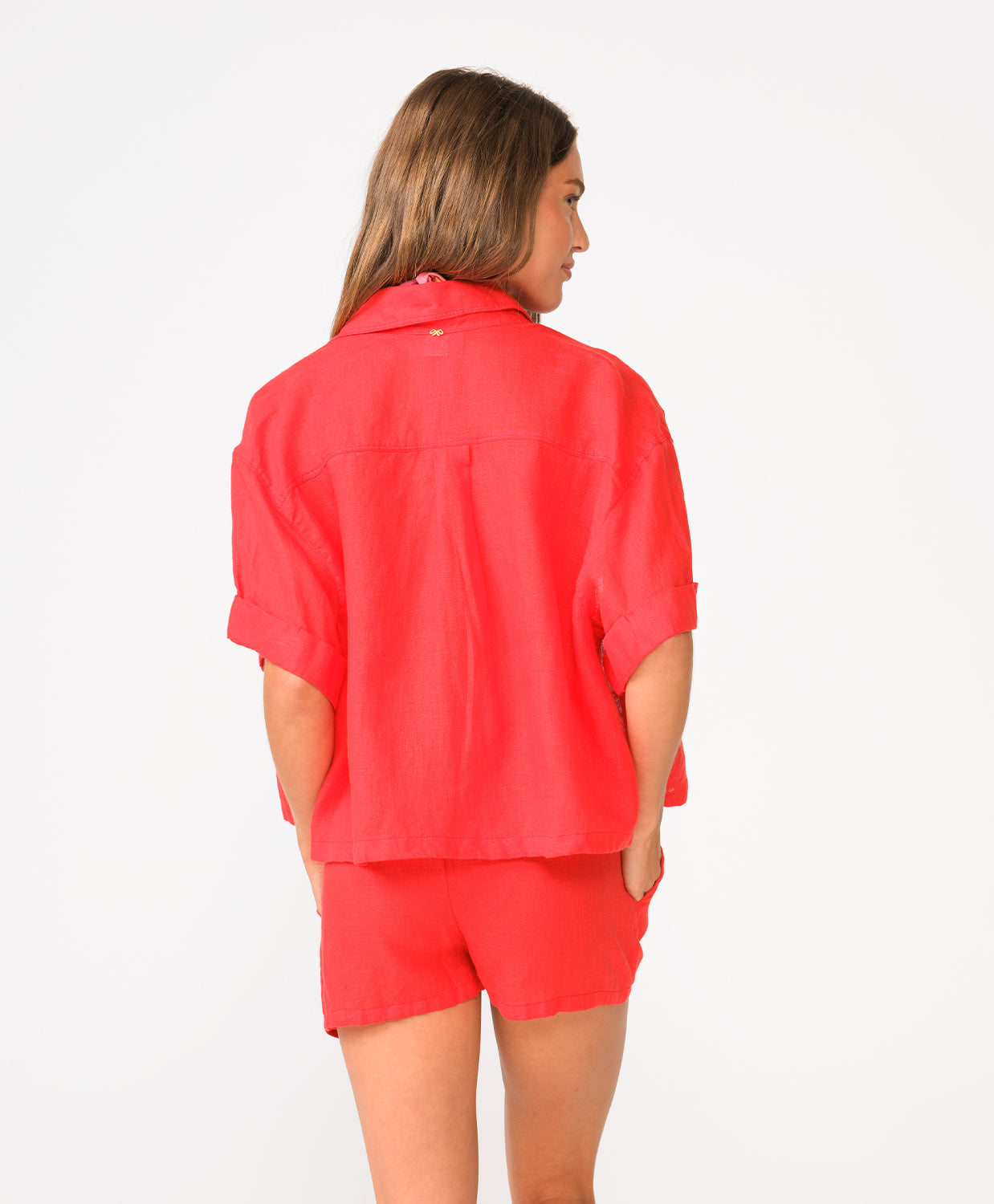 A woman wearing a red short sleeve linen top and matching linen shorts facing towards a white wall. 