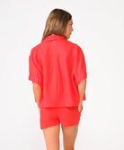 A woman wearing a red short sleeve linen top and matching linen shorts facing towards a white wall. 
