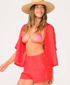 A woman wearing a pink and red bikini top, red short sleeve linen top, and matching linen shorts standing against a white wall. 