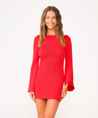 A woman wearing a long sleeve red mini dress standing against a white wall. 