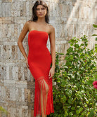 A woman wearing a strapless red mesh dress with fringe standing in front of a concrete wall outside. 