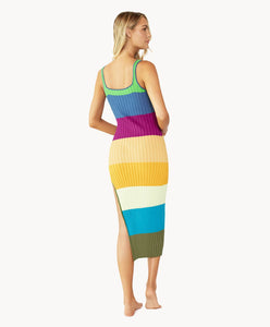 Blonde woman wearing a multi-colored stripe pattern dress with side slit facing backwards towards white wall.