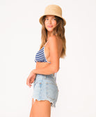 A woman wearing a blue and white stripe triangle bikini top and jean shorts standing against a white wall.
