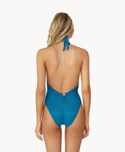 Blonde woman wearing a textured turquoise halter one-piece swimsuit with gold details facing backwards towards a white wall.
