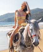 Woman in a colorful bikini sitting on a horse by the beach with mountains in the background