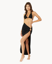Brunette woman wearing a black halter bikini with net macramé details & shimmery gold accents and black skirt coverup with ring detail stands in front of white wall.
