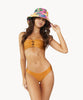 Brunette woman wearing an orange bandeau bikini with gold details and keyhole cutouts and pink tropical pattern reversible bucket hat stands in front of white wall.