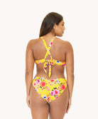 Back view of brunette woman wearing a yellow and flower-patterned bikini in front of a white background. 