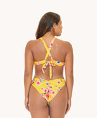 Back view of brunette woman wearing a yellow and flower-patterned bikini in front of a white background. 