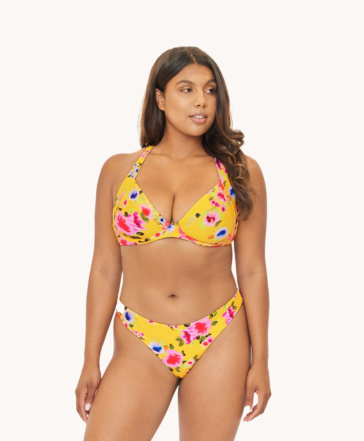 Brunette woman wearing a yellow and flower-patterned bikini in front of a white background. 