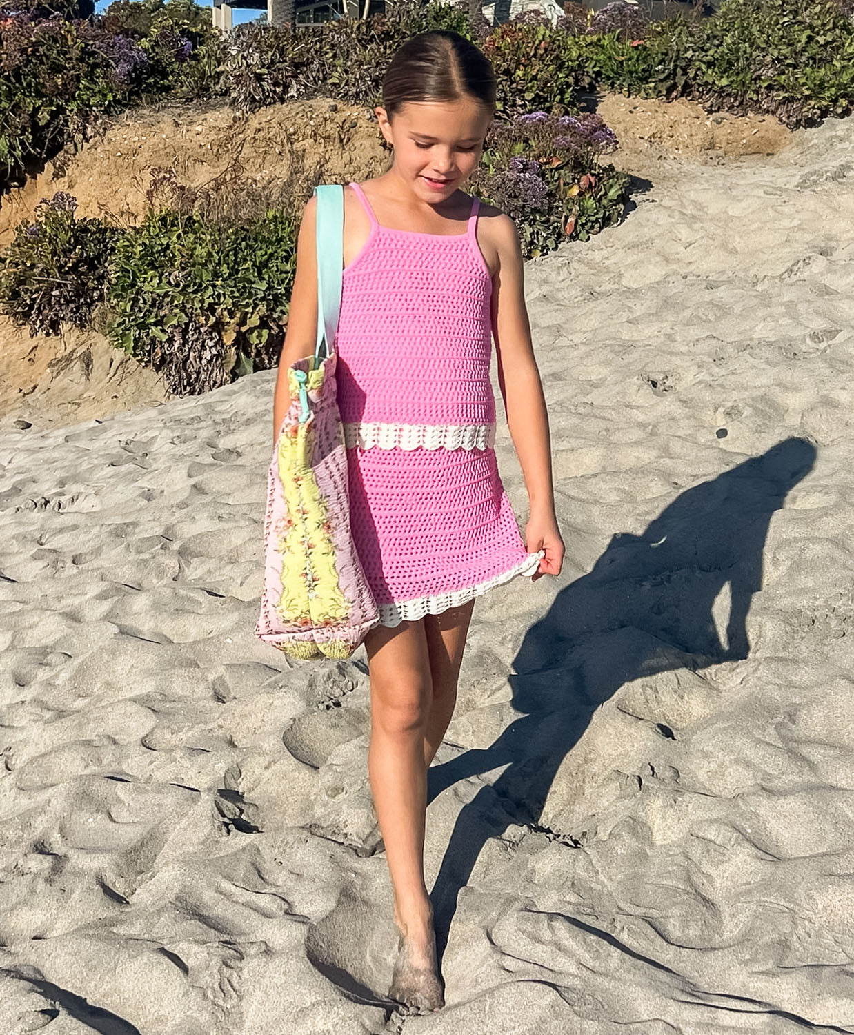 Young girl in a pink dress walking on a sandy beach with a floral bag.
