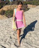 Young girl in a pink dress walking on a sandy beach with a floral bag.
