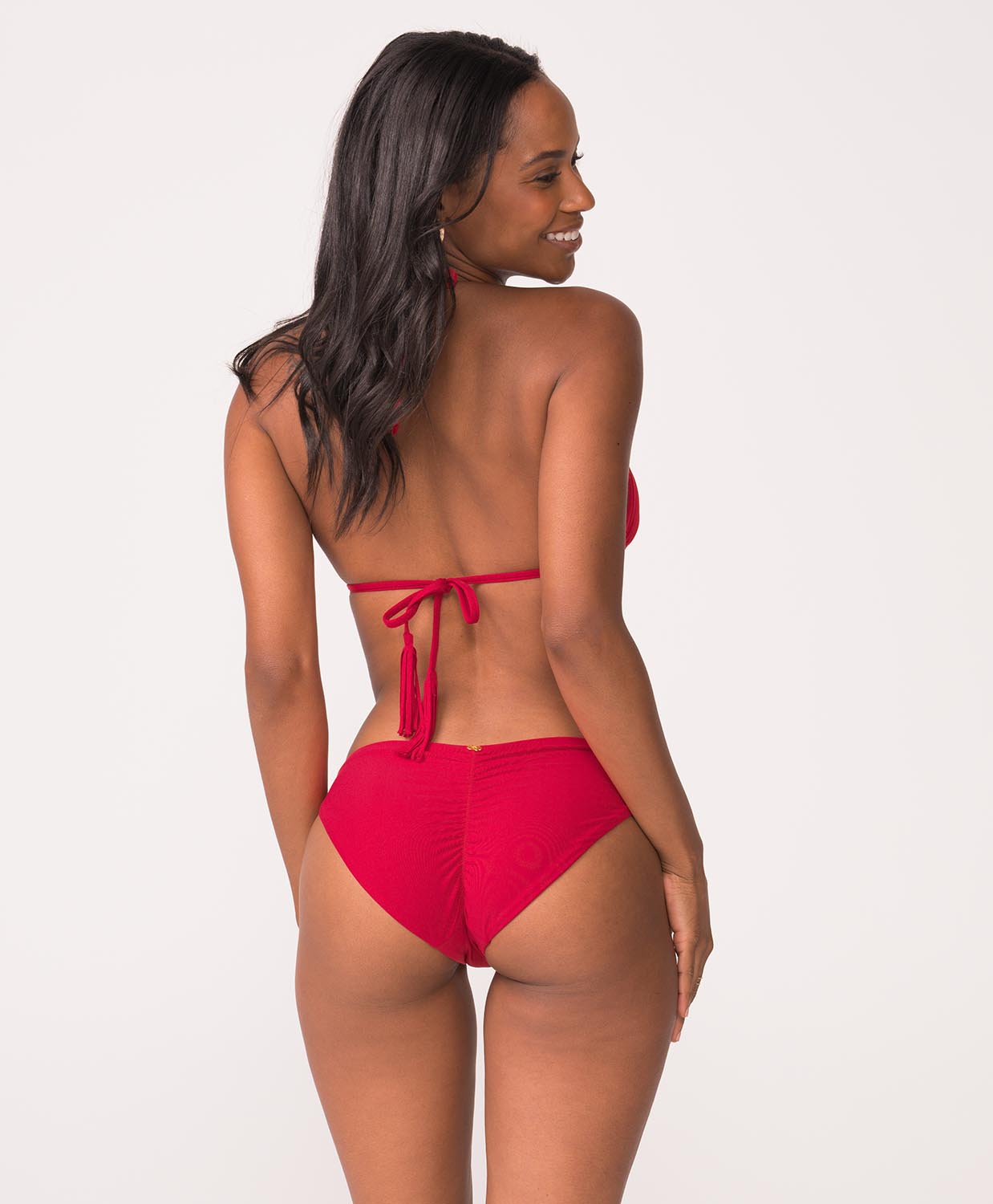 Woman wearing a red bikini with ruched bottoms stands in front of white wall.