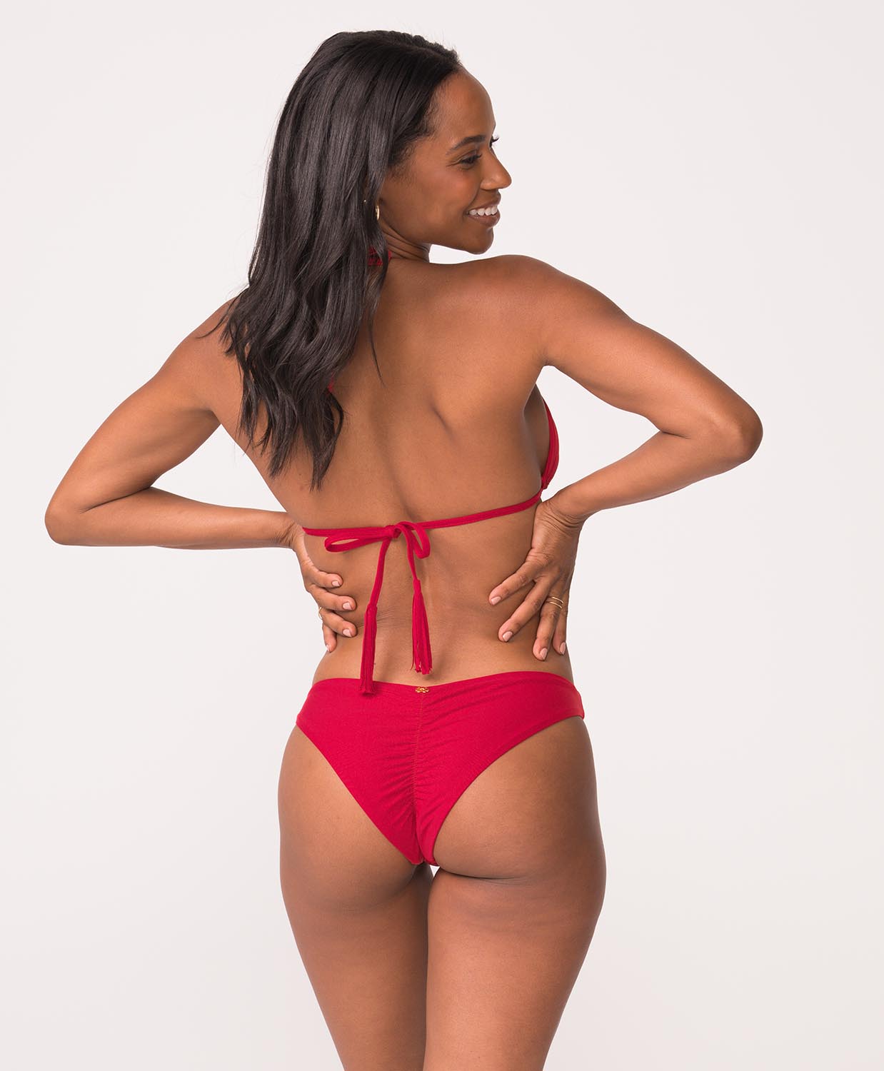 Woman wearing a red bikini with ruched bottoms stands in front of white wall.