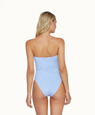 Back view of blonde woman wearing a stripped white and blue one piece in front of a white background. 