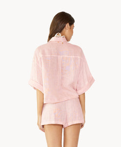 Brunette woman wearing a pink linen button coverup shirt and shorts facing backwards towards white wall.