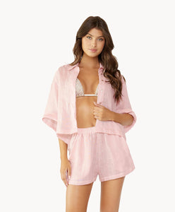 Brunette woman wearing a triangle bikini top under a pink linen button coverup shirt and shorts stands in front of white wall.
