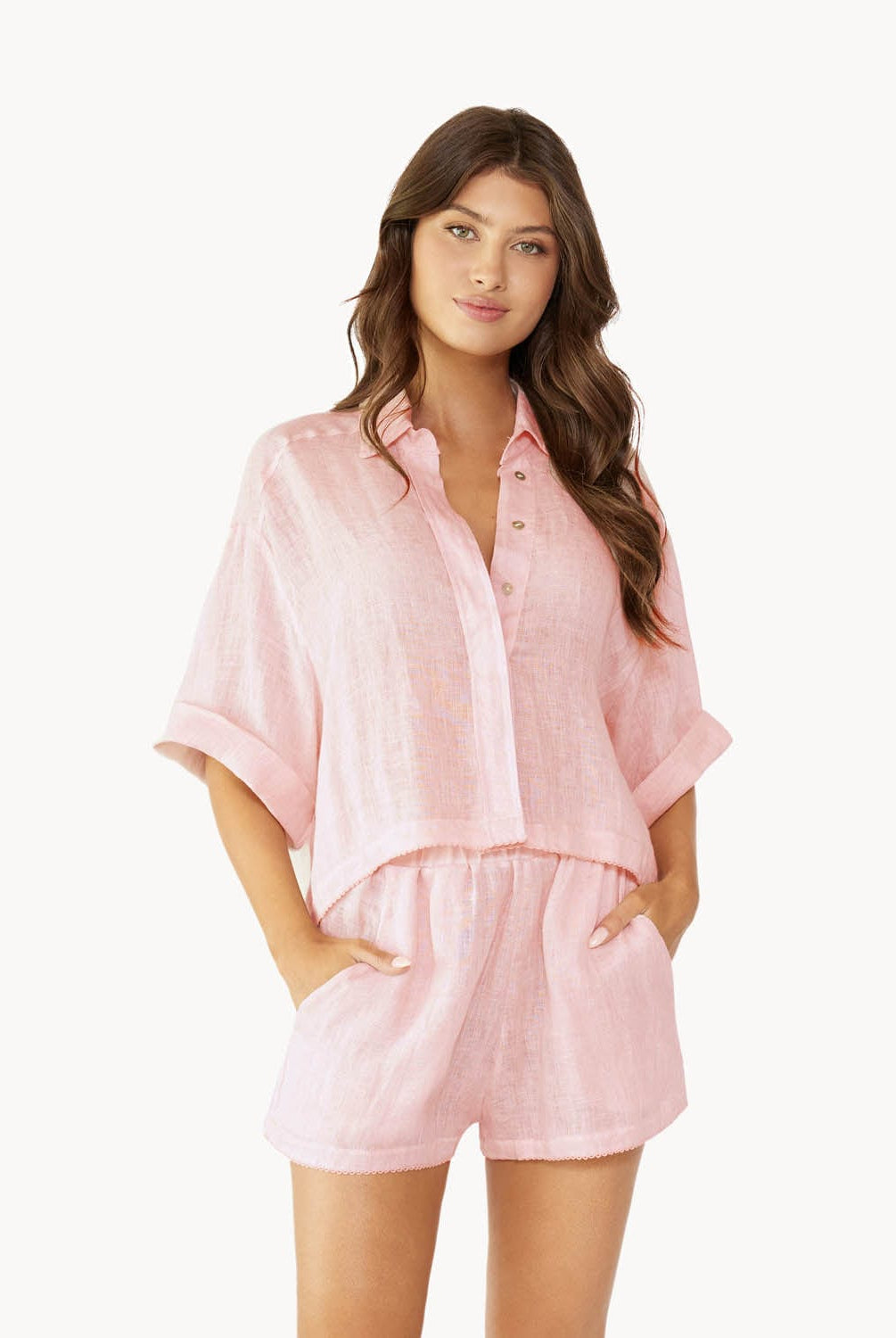 Brunette woman wearing a pink linen button coverup shirt and shorts stands in front of white wall.