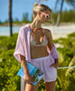 Blonde woman wearing a pink linen coverup shirt and shorts with a multi-colored print triangle bikini with gold details stands by the beach.