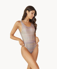 Brunette woman wearing a multi-colored print one piece swimsuit stands in front of white wall.